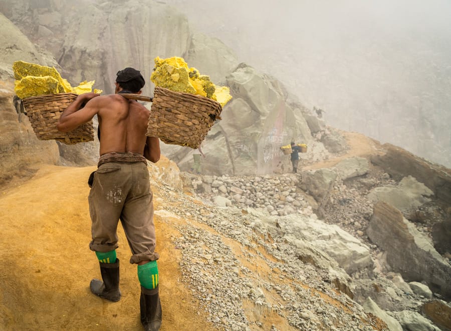 A man climbing the side of a steep mountain slope, hauling two baskets of minerals on his shoulder